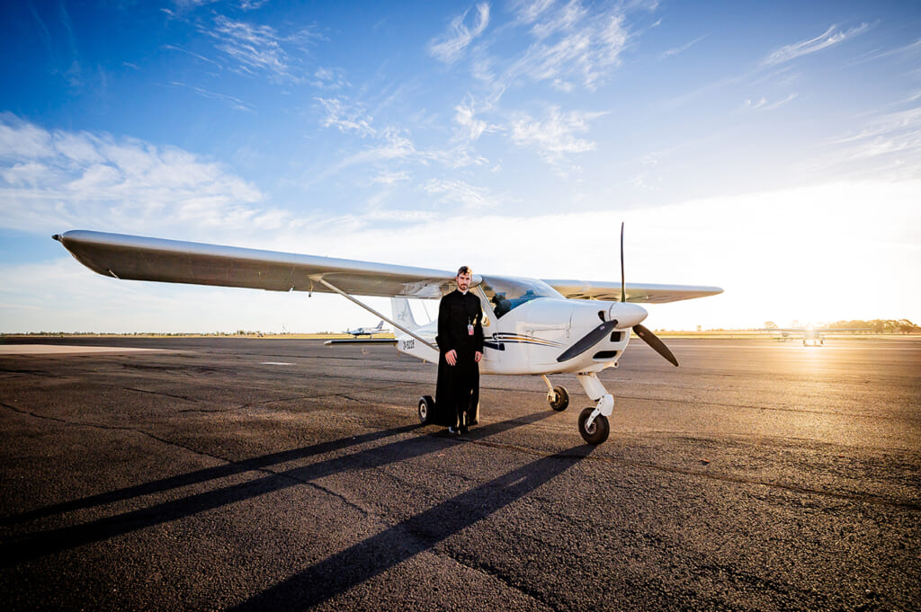 A priest dressed in a black soutane standing by a single person light areoplane parked on a runway. The sun is setting in the background.